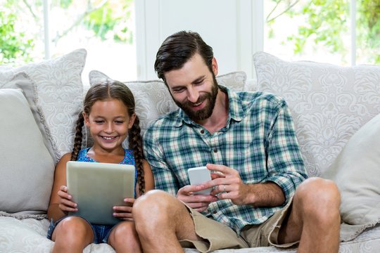 Playful daughter and father with digital tablet and mobile sitting on sofa