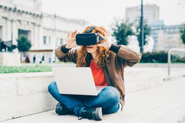 Young beautiful redhead caucasian business woman  sitting with laptop on his knee using 3D viewer - futuristic, multitasking, technology concept