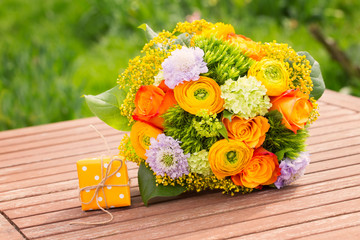 Wunderschöner Blumenstrauß mit Rosen und Ranunkeln und kleines Geschenk auf Gartentisch
