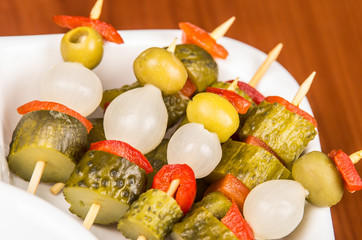 Vegetable skewers of pickles, olives, capiscum and baby onions lying on white plate, wooden surface background as seen from above