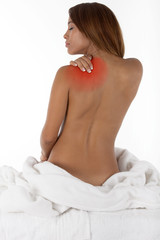 Pretty young woman with white bath robe off sitting on a massage table at a spa waiting for a massage. Rubbing shoulder where its sore, redness added to depict back or shoulder pain.
