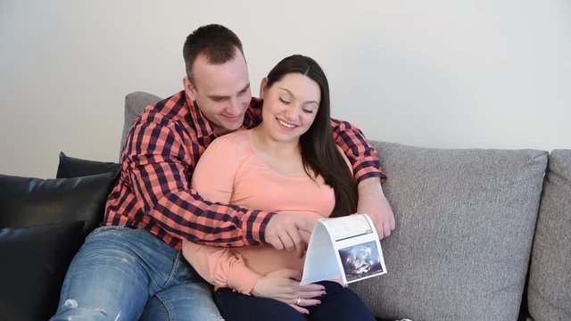 Husband and his pregnant wife sitting on the sofa, smiling, talking and looking at the ultrasound pregnancy photo 