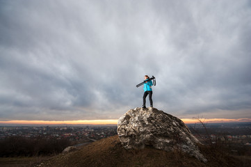 Young attractive female photographer is standing with her camera on tripod on the big rock at city overview point at the sunset. Girl is holding tripod on her shoulder. Blue clouds in the background