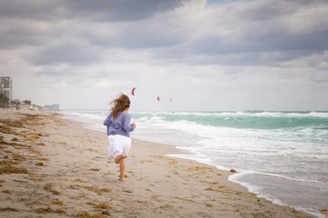 Fototapeta na wymiar Little girl running on the ocean beach on a cloudy day. Vacation by the sea. Cute kid girl on the deserted beach. Summer, outdoors. Wind in the hair of a small girl. Beach landscape.