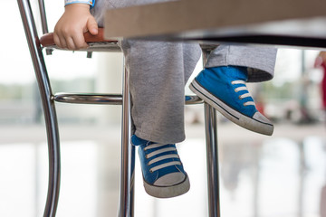Close-up of toddler's legs in blues sneakers under the table. Toddler boy sitting on the high chair...