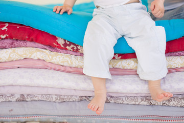 Small baby boy in white trousers sitting on a pile of colorful blankets and mattresses. Happy childhood. Leisure. Indoors. barefoot boy in the bedroom or kids room.