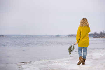 Beautiful blond woman in a yellow sweater and blue jeans walking on the river shore. Happy girl walking on the sand beach. Spring in the city. Enjoying walking outdoors. Snow on the shore.