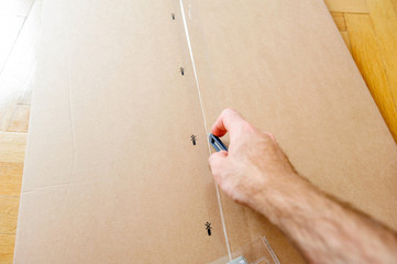 Caucasian man cutting a cardboard box with sharp steel box cutter knife  while installing new furniture