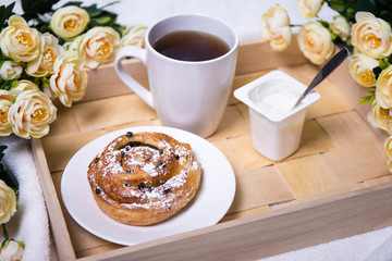 breakfast with bun, tea, yoghurt and flowers on wooden tray