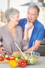 Happy senior woman feeding husband while standing at counter