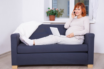 young pregnant woman using laptop and talking by phone at home