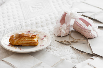 Bitten eclair with cream on plate and pink cute baby booties among paper letters on bed.