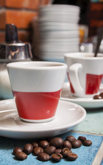 Roasted coffee beans in front of two red and white cup with saucer on the bar - table with blue ceramic tiles.