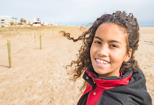 Adorable young girl wearing red coat, walking on beach in Spain in winter. Beautiful brazilian child with long curly hair enjoying wind and sun outside, image for children concept family blog. 