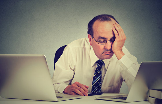 tired sleepy middle aged man sitting at desk with books in front of two laptop computers