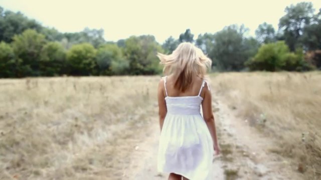 Beautiful young girl with long blond hare happily running along country road on warm summer day. Pretty woman enjoying nature outside.