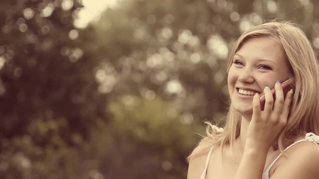 Beautiful young girl having cheerful conversation on cell phone outside. Close up portrait of beauty face of laughing girl talking phone at green trees background. Slow motion video footage.
