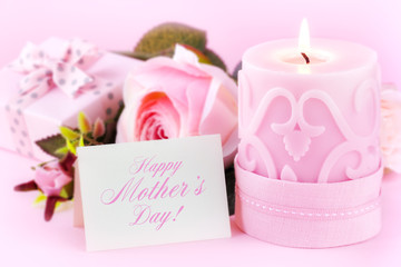 Beautiful festive setting with a greeting card, lit candle, roses and a gift against pastel pink background. With copy space for your text