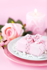 Table place setting with a gift on a plate with roses and candle against pink background