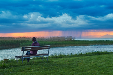 Woman sitting on the bench on the lake coast before an oncoming thunderstorm