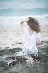 back. Girl with wavy hair on the beach with black sand in a light white top and denim varieties