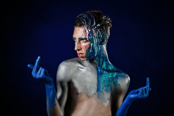 Fototapeta na wymiar Crazy young androgyne man with face art. Freak person. Sperm on face. Tinsel creative blue makeup. Intersex and homosexuality concept. Space for logo. Middle finger 