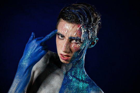 Crazy young androgyne man with face art. Freak person. Sperm on face. Tinsel creative blue makeup. Intersex and homosexuality concept. Space for logo. Middle finger 