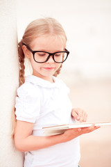 Baby girl 4-5 year old holding tablet computer outdoors. Wearing glasses. Childhood.