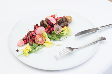 Vinaigrette Russian Salad made of chopped beets, carrots, onion, beans, peas, seasoned with sunflower seed oil, with slices of tomatoes, herbs and sausage, served on a white table with fork and knife