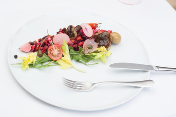 Vinaigrette Russian Salad made of chopped beets, carrots, onion, beans, peas, seasoned with sunflower seed oil, with slices of tomatoes, herbs and sausage, served on a white table with fork and knife