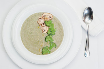 Mushrooms Cream Soup with Pieces of Broccoli and Slices of Mushrooms served in a white plate with a metallic spoon
