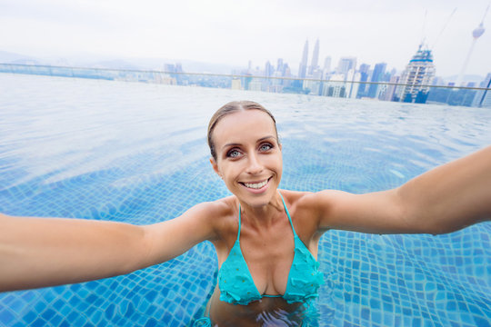 Vacation and technology. Pretty young woman taking selfie while swimming in roof top pool with beautiful city view.