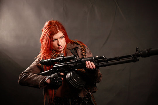 Studio shot of attractive redhead punk girl with rifle and sniper equipment, taken againstwall covered with dark fabric