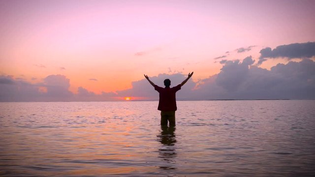 Man raises his arms to a worship pose in a calm ocean to a morning surise