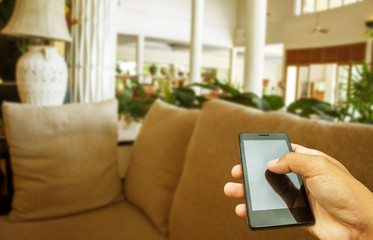 Hand hold and touch screen smart phone, tablet,cellphone on blur image of modern living room at home.