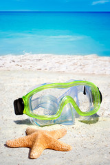 Fototapeta na wymiar Taucherbrille und Seestern liegen im Sand am Meeresstrand mit Wellen im Hintergrund
