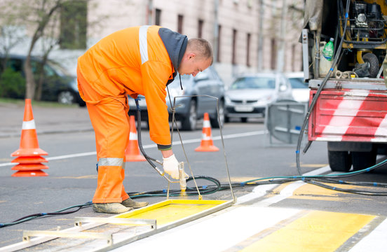 Technical person man worker painting and remarking pedestrian crossing lines on asphalt surface using paint sprayer gun during capital street pavement maintenance works