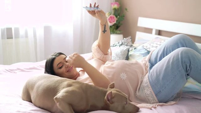 Pregnant woman and pet dog lying on bed and posing for selfie. 4k
