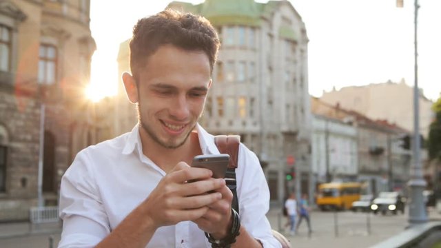 Attractive male in his 20s typing a message on his smartphone in a sunset city center.