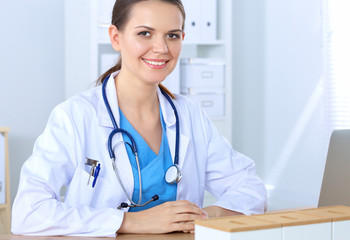 Beautiful young smiling female doctor sitting at the desk and writing.