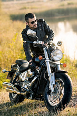 Obraz premium young brutal man in a black jacket and glasses standing near a motorcycle