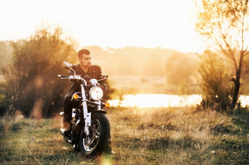 young brutal man in a black jacket and glasses sits near a motorcycle.