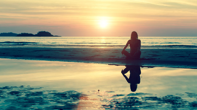 Silhouette of young girl in yoga pose sitting on the beach during amazing sunset.