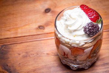 Traditional English dessert strawberry trifle with blackberries in a transparent glass on a rustic wooden surface. Free space for text