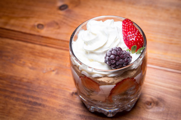 Traditional English dessert strawberry trifle with blackberries in a transparent glass on a rustic wooden surface. Free space for text