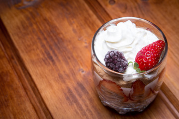 Traditional English dessert strawberry trifle with blackberries in a transparent glass on a rustic wooden surface. Free space for text
