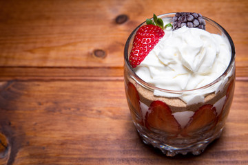 Traditional English dessert strawberry trifle with blackberries in a transparent glass on a rustic wooden surface. Free space for text