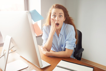 Young female office worker looking surprised and shocked at the camera while working at her desk in front of computer. Business and office concept. Human face expressions and emotions
