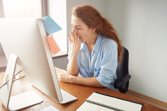 Portrait Of Tired Sleepy Businesswoman Yawning, Working At Desk In The Office In Front Of Computer. Bored Young Female Freelancer Working On PC At Home. Overwork And Sleep Deprivation Concept