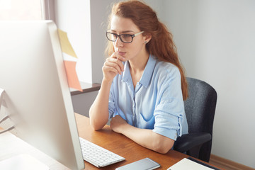 Confident female freelancer sitting in front of the computer with serious and thoughtful expression. Young businesswoman in blue shirt and glasses using PC for work sitting in the office